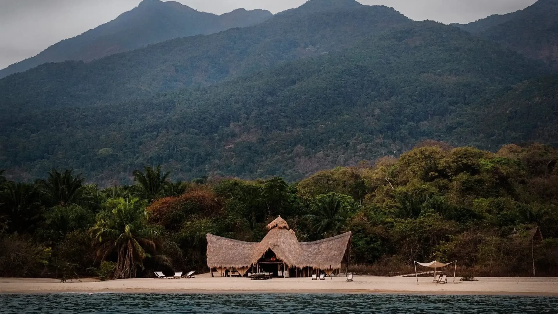 A thatched lodge on a sandy lake shore with forested mountains rising behind it, Greystoke Mahale, Lake Tanganyika